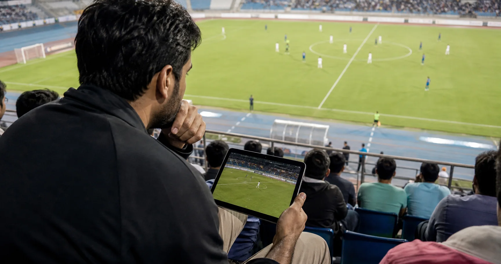 A scout watching an Indian football match from the stands, with football action, pitch markings, and a scouting tablet visible