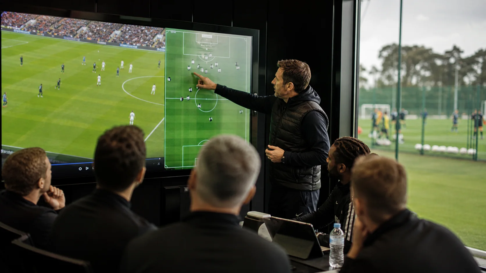A coaching staff briefing in front of a screen that explains a tactical decision using a visible football match frame and pitch map