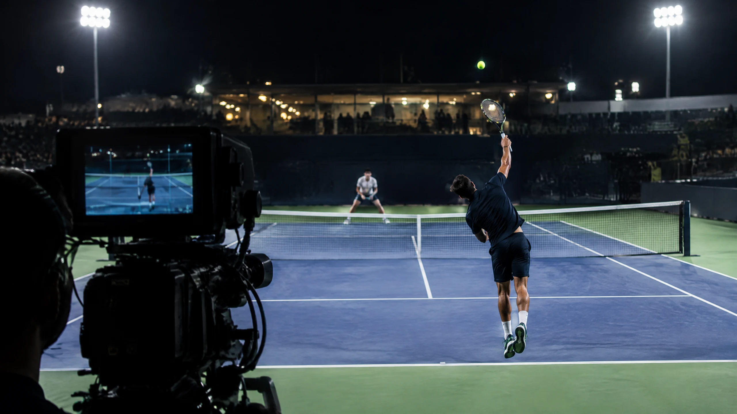 A broadcast camera films a tennis player serving during a night match.