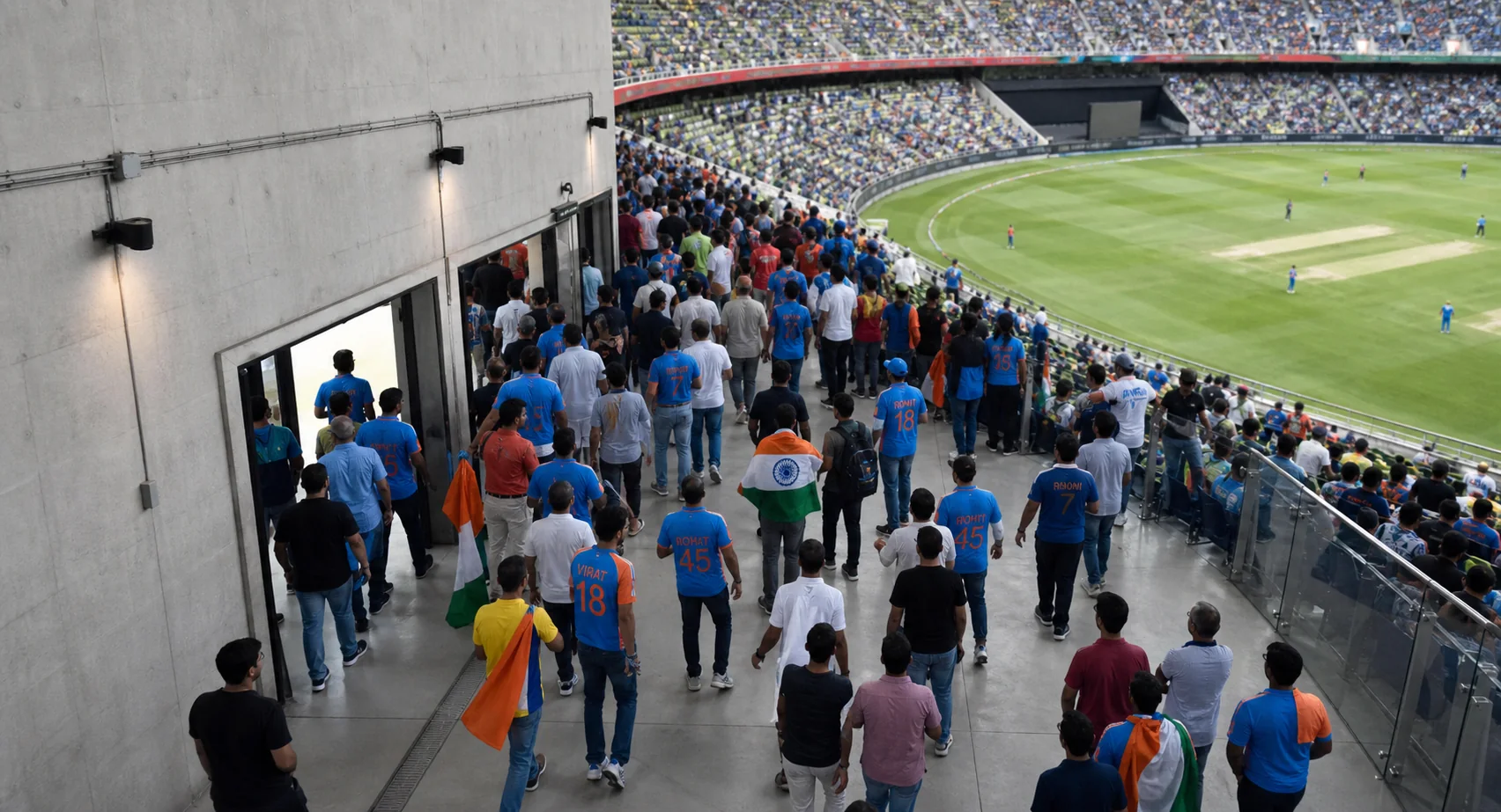 An overhead view of supporters in team apparel moving through a stadium concourse toward a clearly signed entrance, with the football or cricket venue apparent