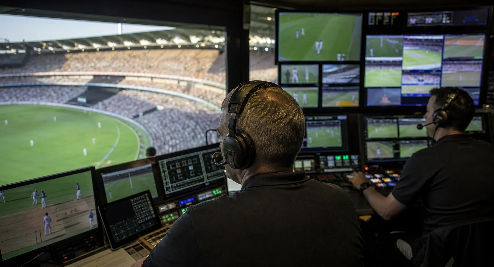 A remote-production control room whose video wall shows multiple camera angles of a live football or cricket match and a clearly recognisable stadium bowl