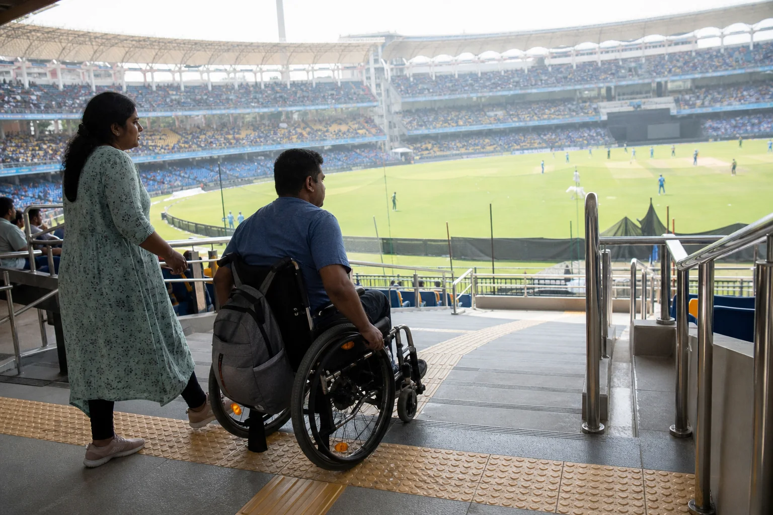 A wheelchair-using supporter and companion following tactile paving and accessible wayfinding inside an Indian stadium, with a visible match field nearby